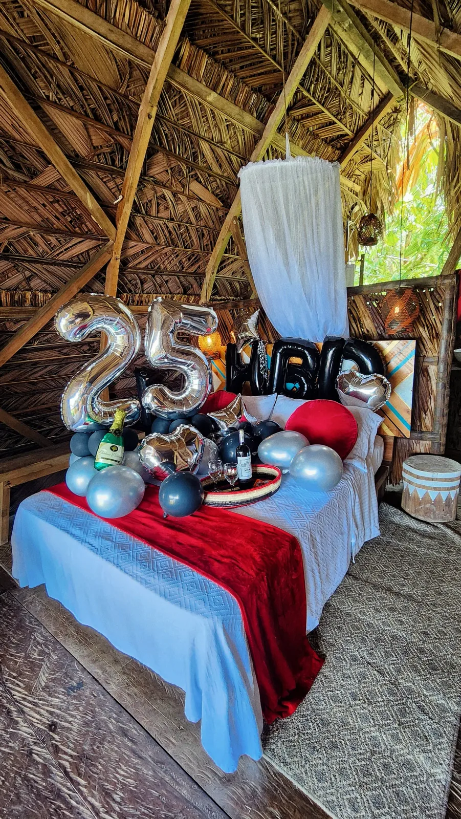 Cama decorada con globos del número 25, pétalos y telas rojas.