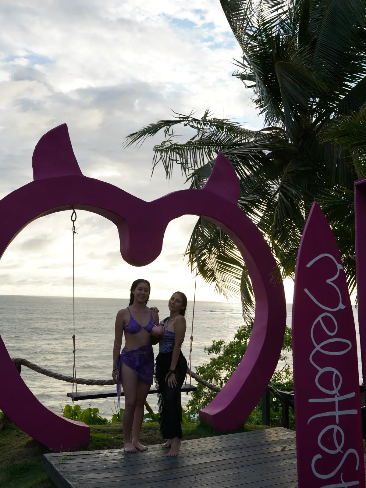 Dos amigas posando en el columpio rosa con forma de corazón frente al mar.