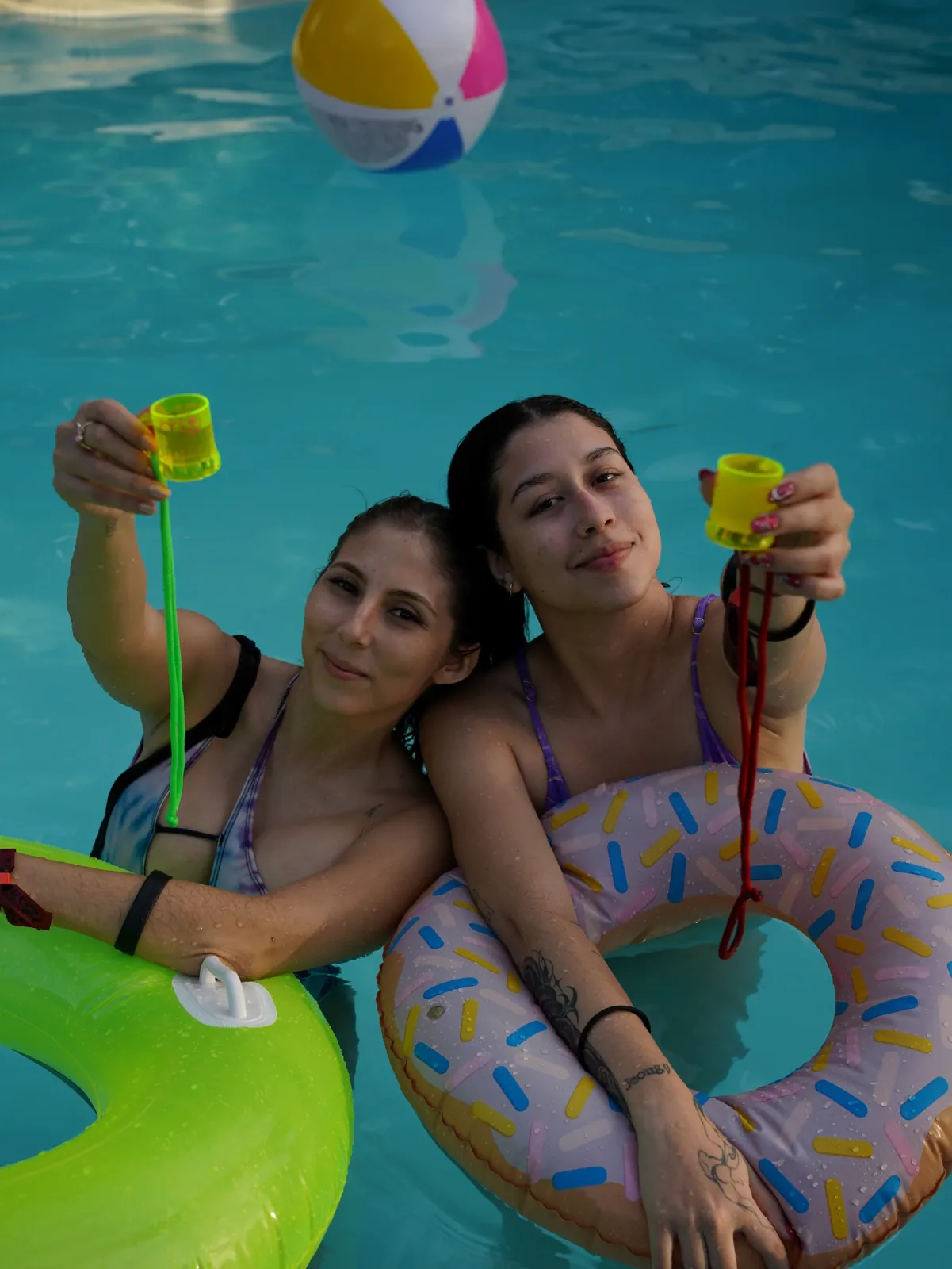 Dos amigas con bebidas y flotadores de colores dentro de la piscina.
