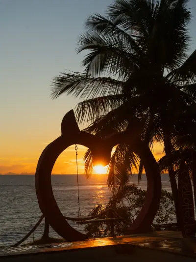 Sol del atardecer enmarcado por palmeras y el columpio con forma de corazón.