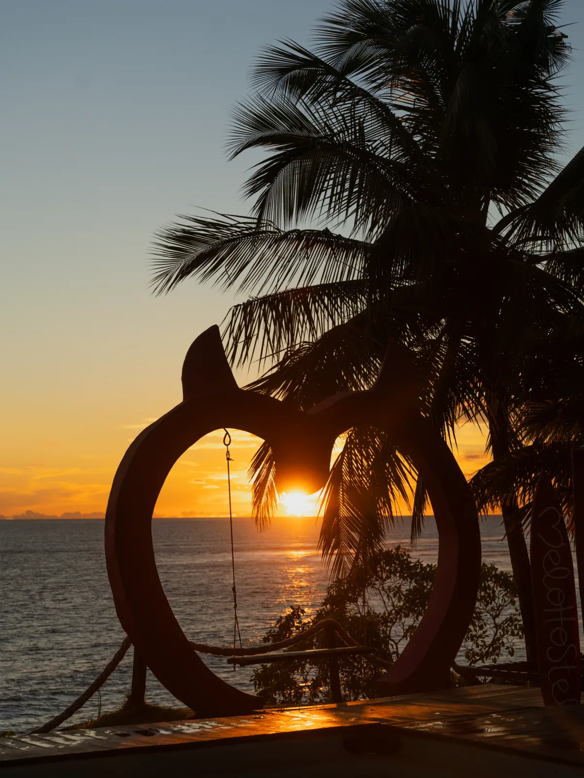 Sol del atardecer enmarcado por palmeras y el columpio con forma de corazón.