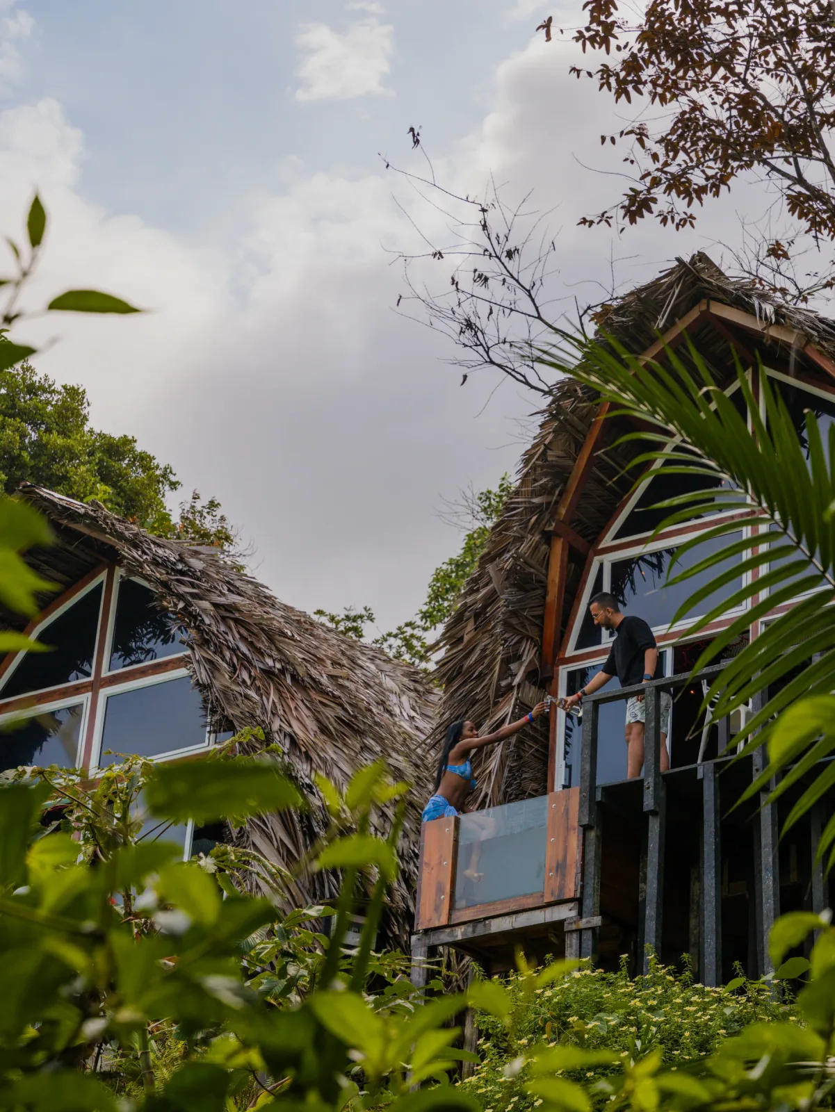 Cabaña triangular elevada con balcón y techo de palma entre la vegetación.