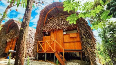 Cabaña tropical de madera con techo de palma entre árboles frutales.