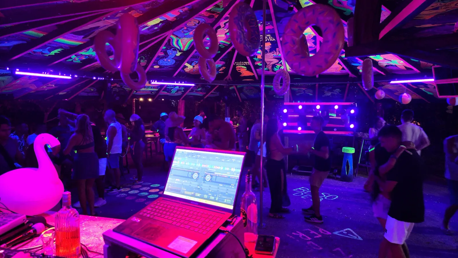 Cabina del DJ en primer plano y público bailando bajo luces neón.