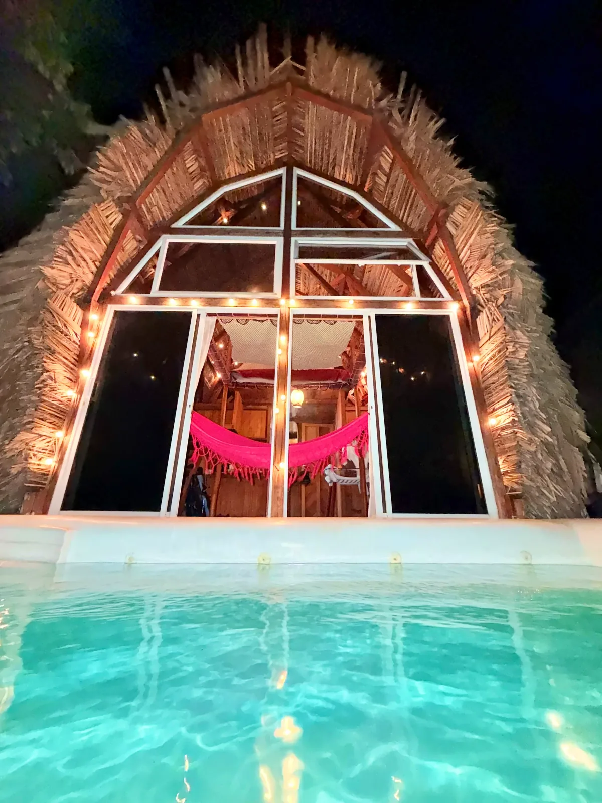 Fachada de Mamámelo iluminada de noche junto a la piscina.