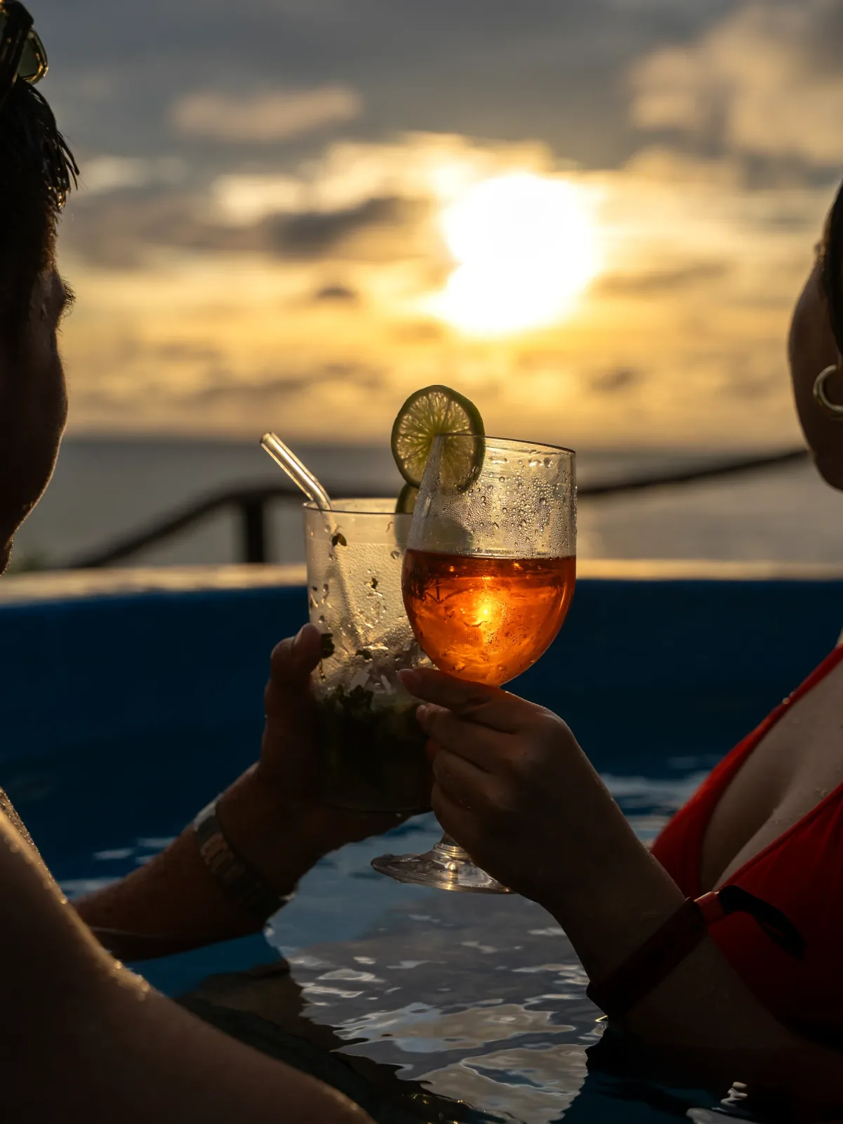 Pareja brindando con cócteles junto a la piscina durante el atardecer.