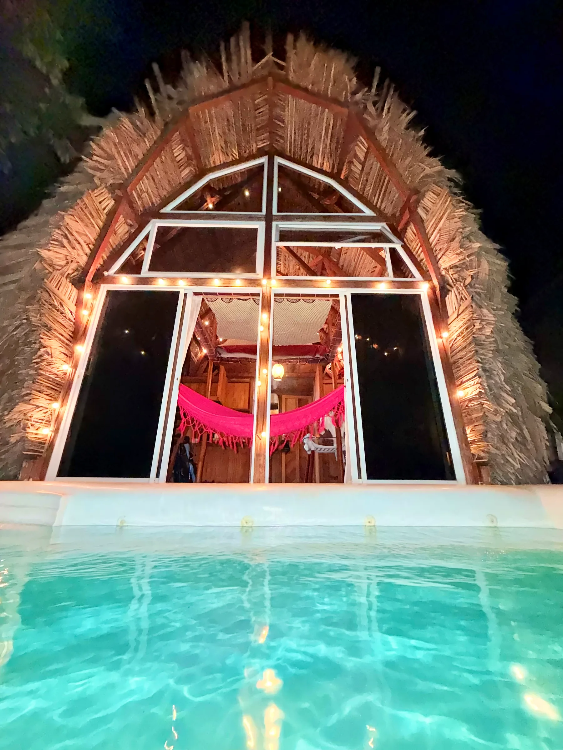 Fachada de Mamámelo iluminada de noche junto a la piscina.
