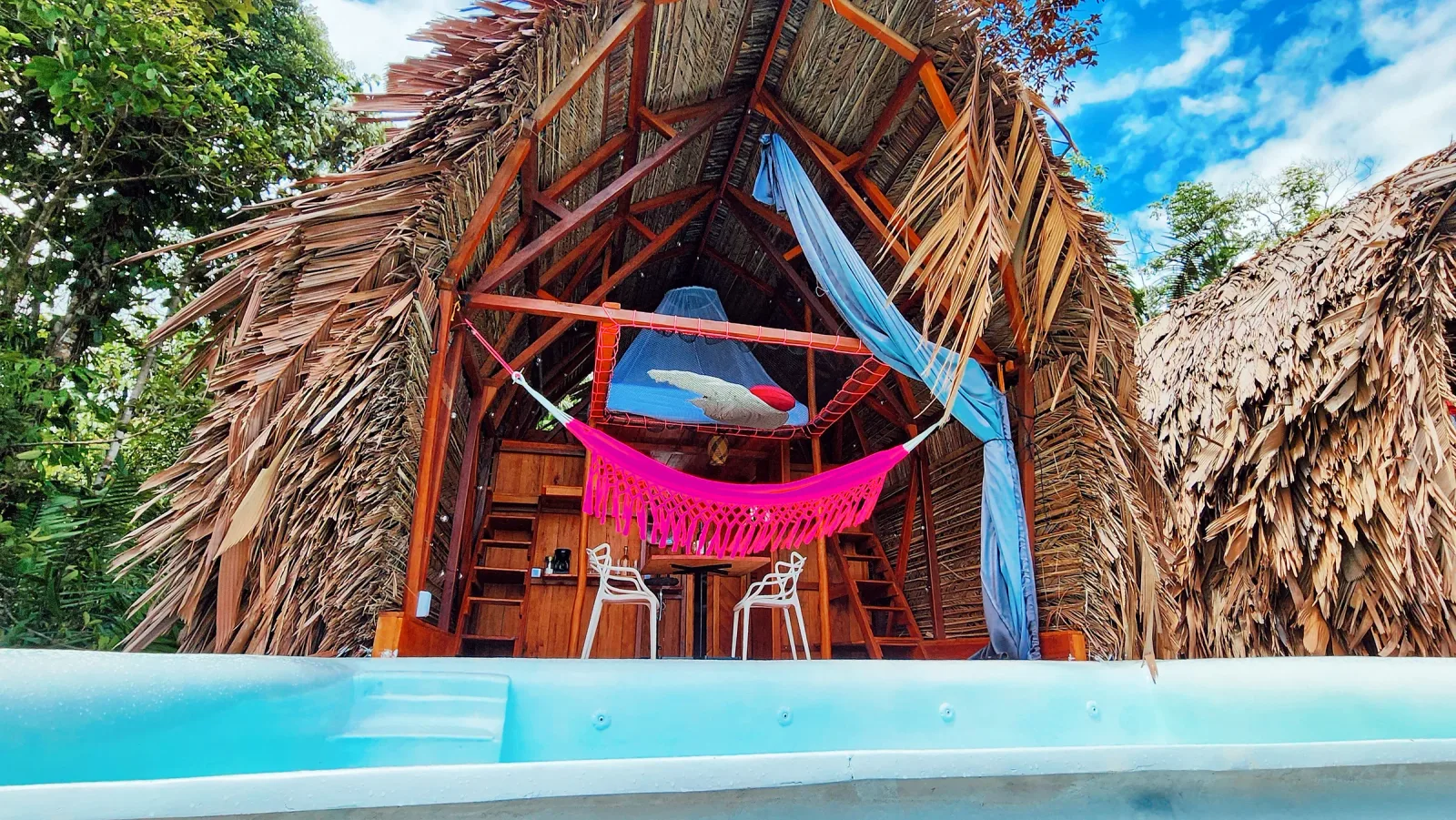 Cabaña de madera con techo de palma, hamaca rosa y piscina privada.